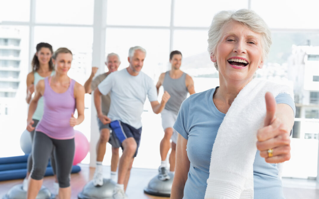 Cheerful senior woman gesturing thumbs up with people exercising in the background at fitness studio